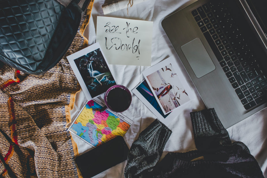 Flatlay of a mug of coffee, photos, a small world map, paper with writing that says See The World, all fit next to a cellphone and laptop.