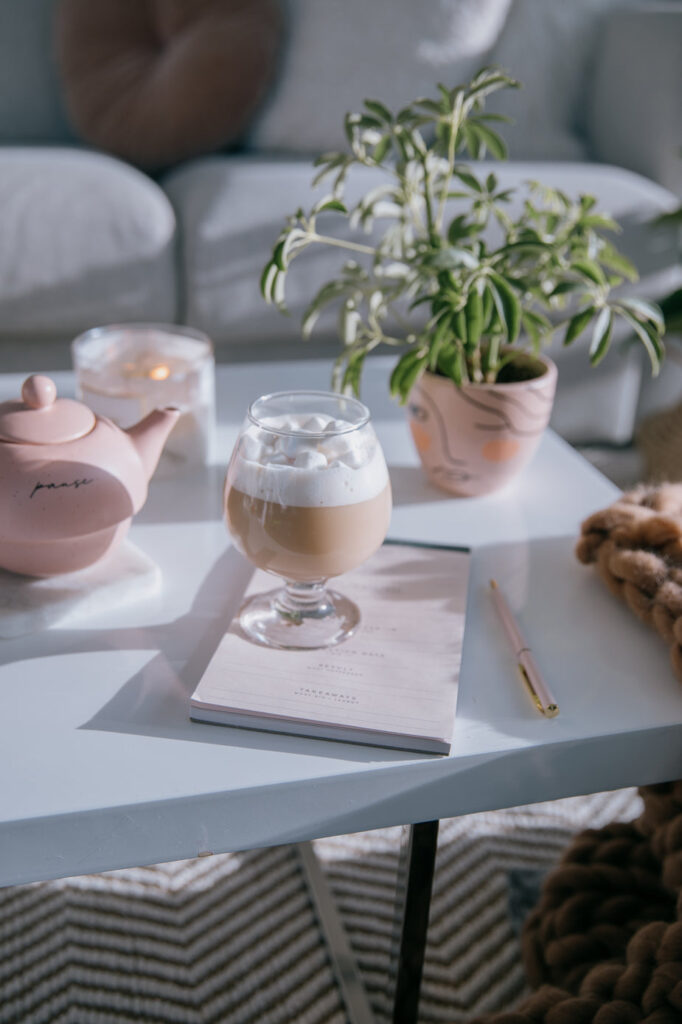 A white coffee table with a teapot, a lit candle, a house plant in a pink pot and a round glass with hot chocolate topped with marshmallows sits on top of a pad of paper with a pen laying next to it. A couch is out of focus in the background. Student teaches themselves to become a scopist