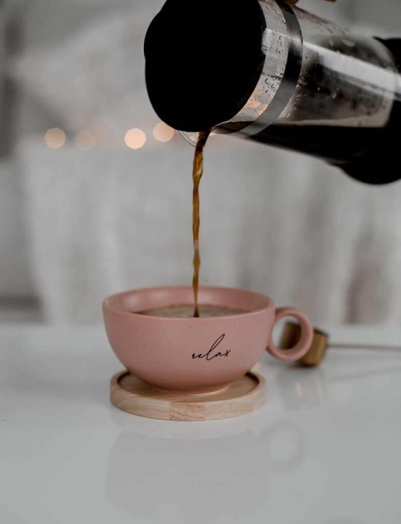 A french press is pouring black coffee into a pink coffee cup, that has the word Relax on it. The cup is sitting on a light wooden coaster on a white table while taking a break from learning to be successful