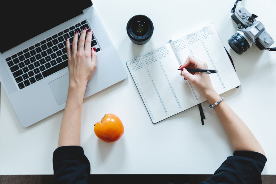 Top view of a white desk with a person with one hand on a laptop keyboard and the other writing in a open notebook. There is a laptop, camera, a persimmon and camera lens sitting on the table as well.