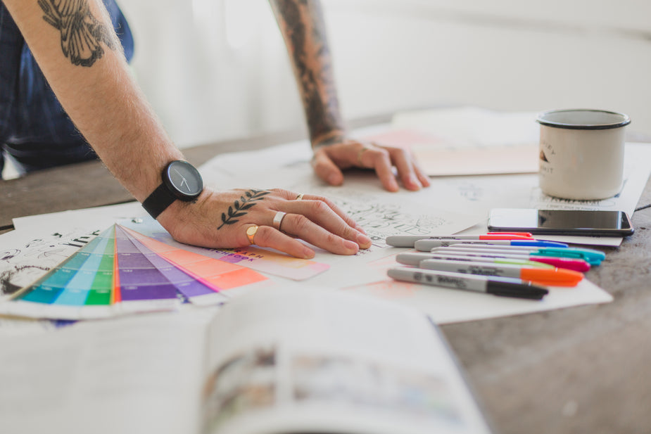 A view of the tattooed arms of a man leaning against his worktable. There are color swatches, markers, a cup of coffee, and potential designs that fit spread across his desk. 