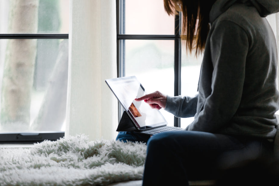 Sitting on a comfy window seat, a woman uses the touch screen of her tablet.