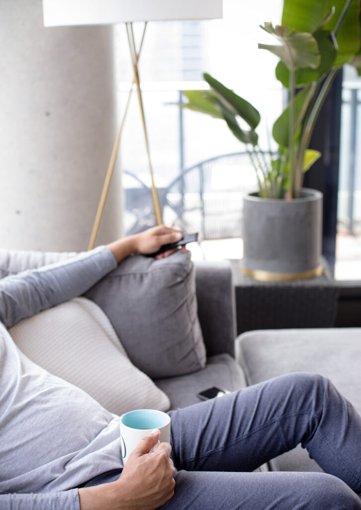 Person holds a mug on their lap and sits comfortably on a grey couch. Out of focus you can see a large house plant