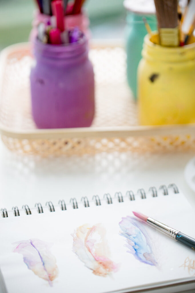 A paint brush lays next to a water color painting of three colorful feathers.