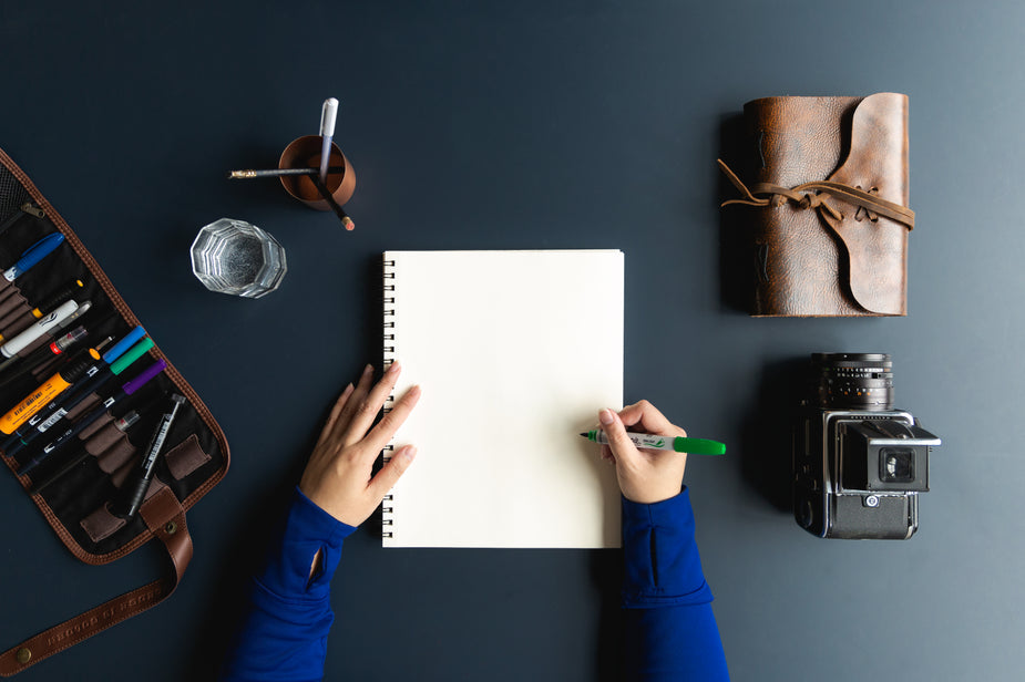 A hand hold a green felt tipped pen ready to create content for building an online business on the blank pages of a notebook. Surrounded by creative tools such as camera, pencils, journal and a coffee, this artist is ready!