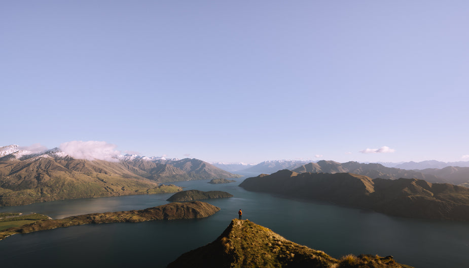 A hiker stands on a mountain peak overlooking a vast mountain landscape, a blue lake dividing it all up. The long journey was worth this breathtaking view.