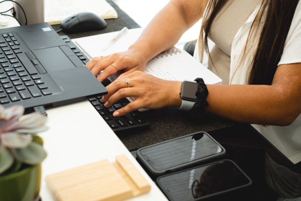 View of a scopist typing on a keyboard at their desk. You can see two cell phones, two keyboards, and open notebook and a potted plant on the desk as well.