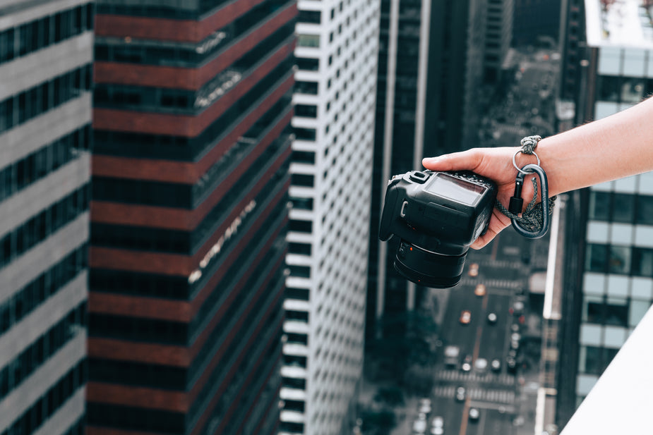 A hand grasping a digital camera reaches over the edge of a building in a city to take a photograph of the busy streetscape below. To ensure it won't fall and hit the subjects below, the camera is affixed to the photographer's wrist by a thick rope and carabiner.