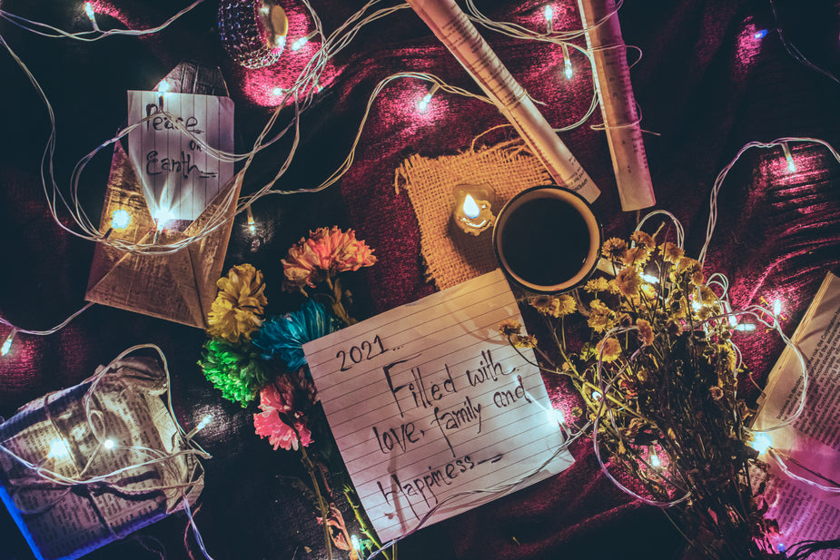 Lined paper with the words '2021 filled with love, family, and happiness' lays on a pink textured cloth surrounded by flowers, string lights, coffee, lit candle, a gift wrapped in newsprint, and a second note that says 'peace on earth'.