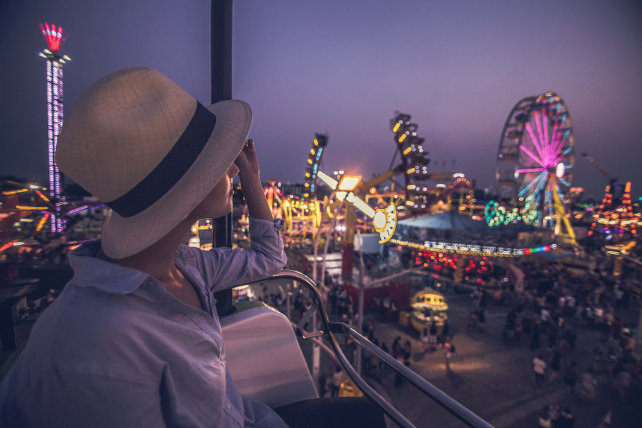 A woman in a summer hat takes a moment to really enjoy the lights and sounds of a carnival at night.