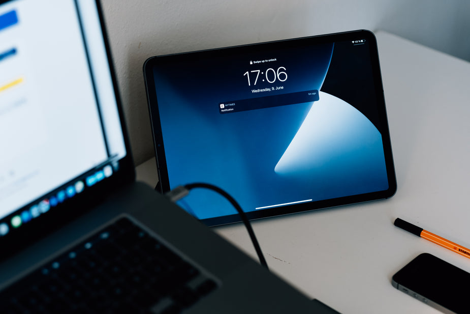 A tablet is leaning against the wall next to a laptop, and above a cellphone. The tablet screen is blue and showing a notification.