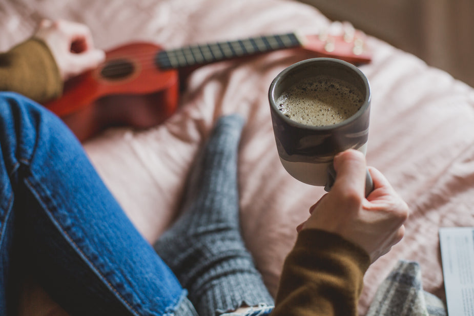 A person sits cross-legged on a bed. In the frame are one leg wearing blue jeans, one foot wearing a sock, and two arms wearing yellow sleeves. The right-hand holds a full cup of coffee, the left hand plucks at a ukelele lying flat on the blanket. Comfort is necessary in your workspace for productivity.