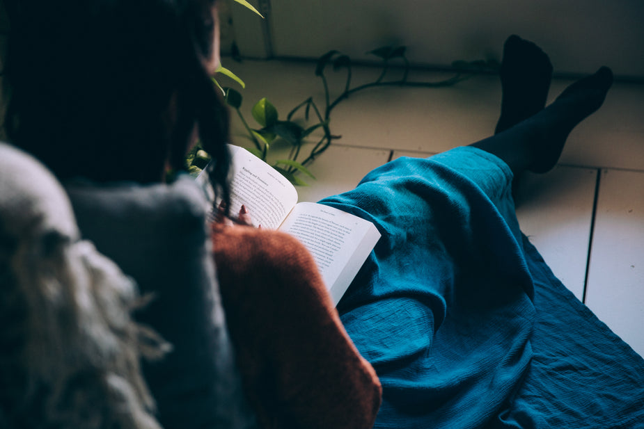 Sitting on a tiled floor, this woman is engrossed in her book. Her long blue shirt blankets her crossed legs and beside her the tendrils of a plant stretch out.