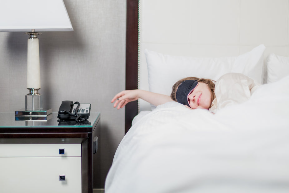 A woman is sleeping with a face mask in a hotel room. A subtle smile could mean a happy dream.