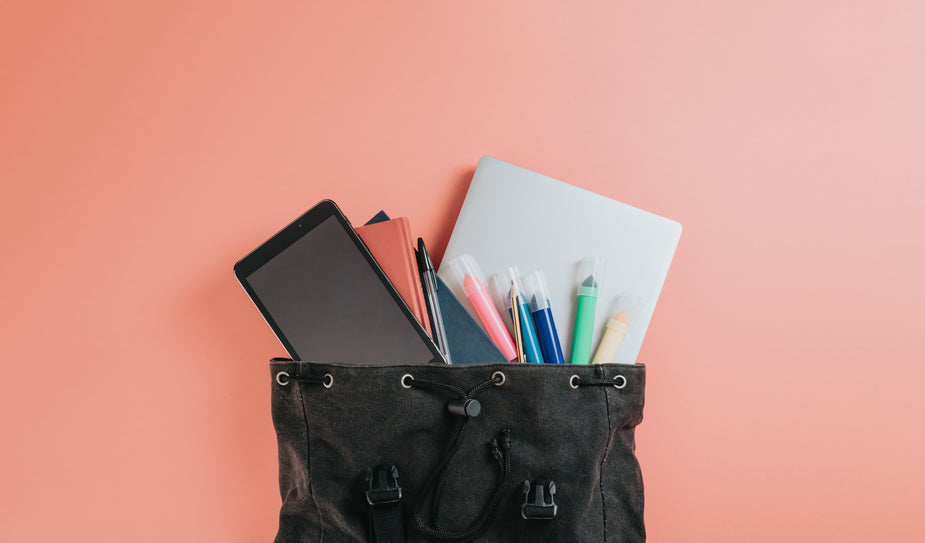 A black bag with school supplies falling out of it lays on a pink background in the middle of the frame. There is a cell phone, writing utensils, books and a laptop. productivity