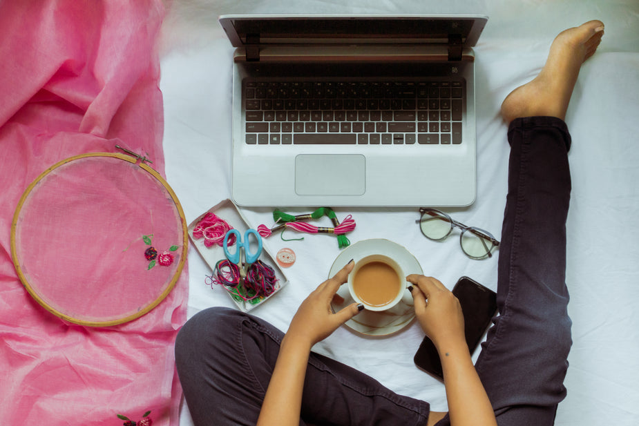 A woman sits on a bed with a tea in hand. Her tools laid out before her to start working at her favorite hobbies. She has her laptop, glasses, phone, and sewing embroidery kit to beat procrastination