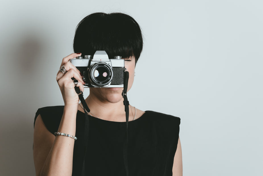 Women with short black hair and black shirt holds a camera up to her face to take a picture.