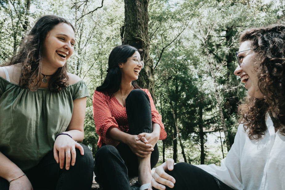 Three people sit together under a large tree in a forest. All three people are laughing and smiling wide to beat procrastination