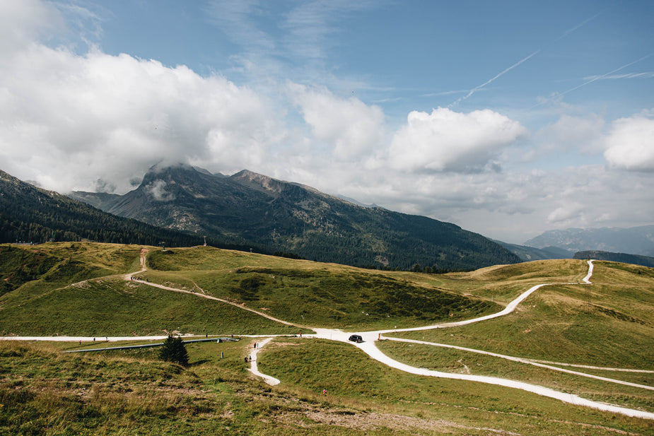 Beneath foggy mountain peaks, a black van sits at an empty intersection of many winding paths. White trails venture off in every direction both on the rolling green hills and the blue clouded sky.