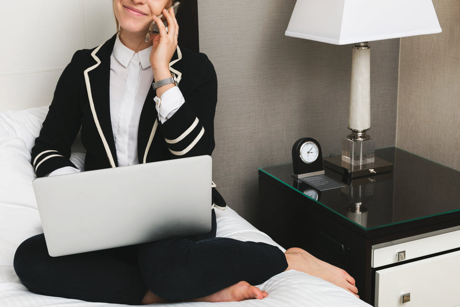 A woman wearing a suit handles a business deal using her phone and laptop while sitting barefoot on her hotel bed. This is a relaxing and productive working vacation! productivity 