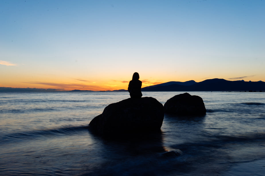 A person takes a moment to reflect and pause as they soak in the last glow of light from an oceanside sunset. The rock they sit on is surrounded by calm waves as the tide rolls in on the beach