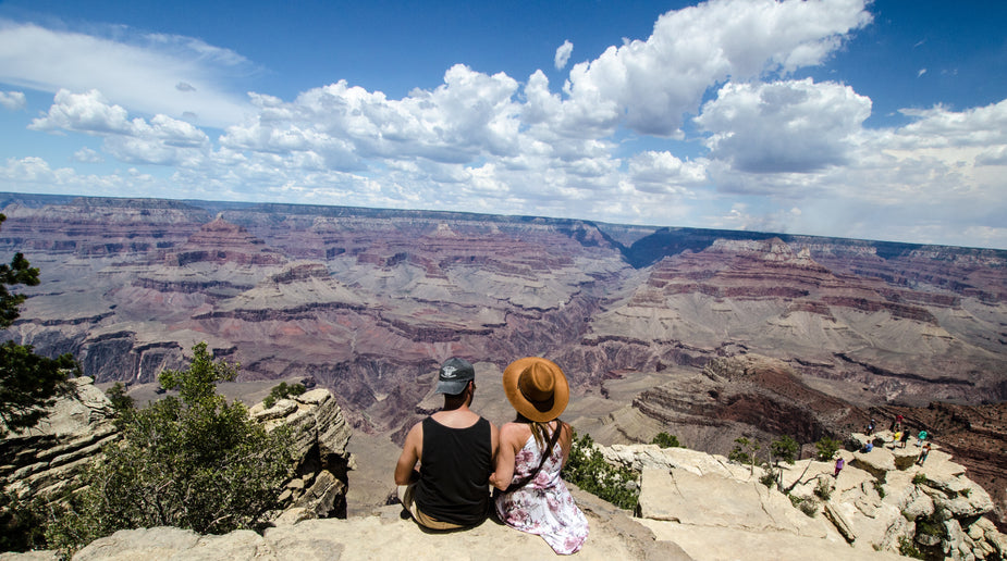 A young man and woman sit on a rocky outcrop overlooking a vast canyon under a blue sky, possibly chatting about life, nature and freedom.