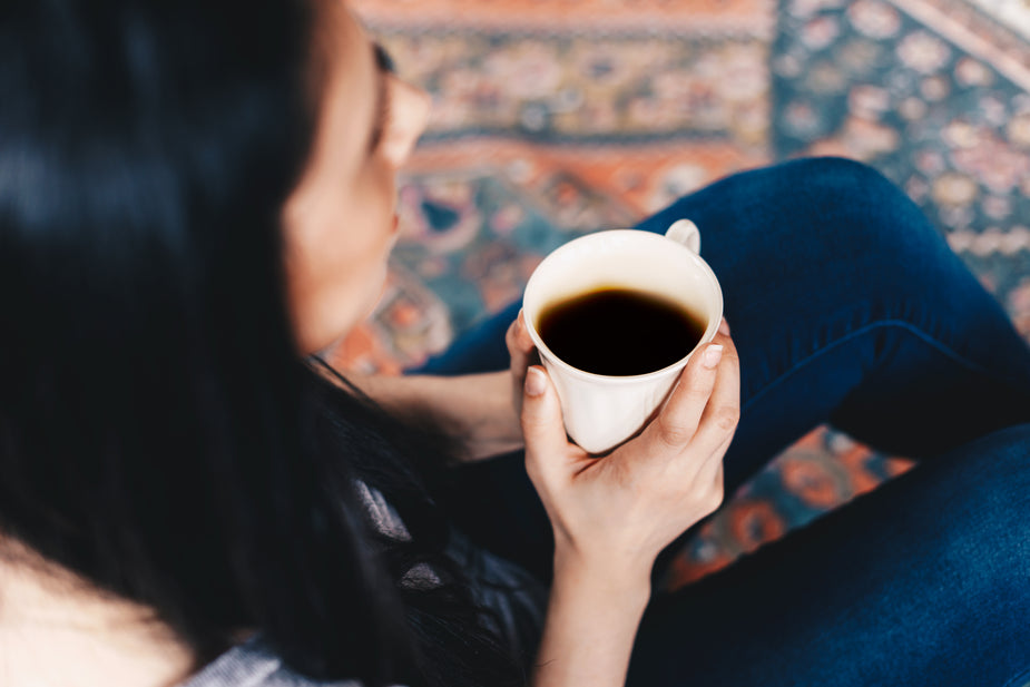 A woman sits on the floor enjoying some peace while drinking a cup of black coffee.