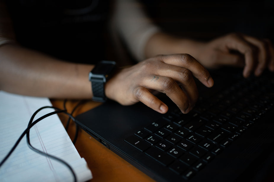 Close up of a person typing on a black laptop keyboard. The person is wearing a black smart watch and there is white lined paper beside them. scoofer